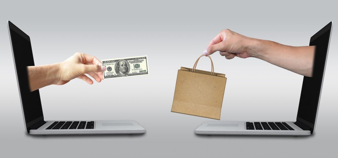 hands reaching out from laptops facing each other one with a hundred dollar bill USD, the other with a shopping bag